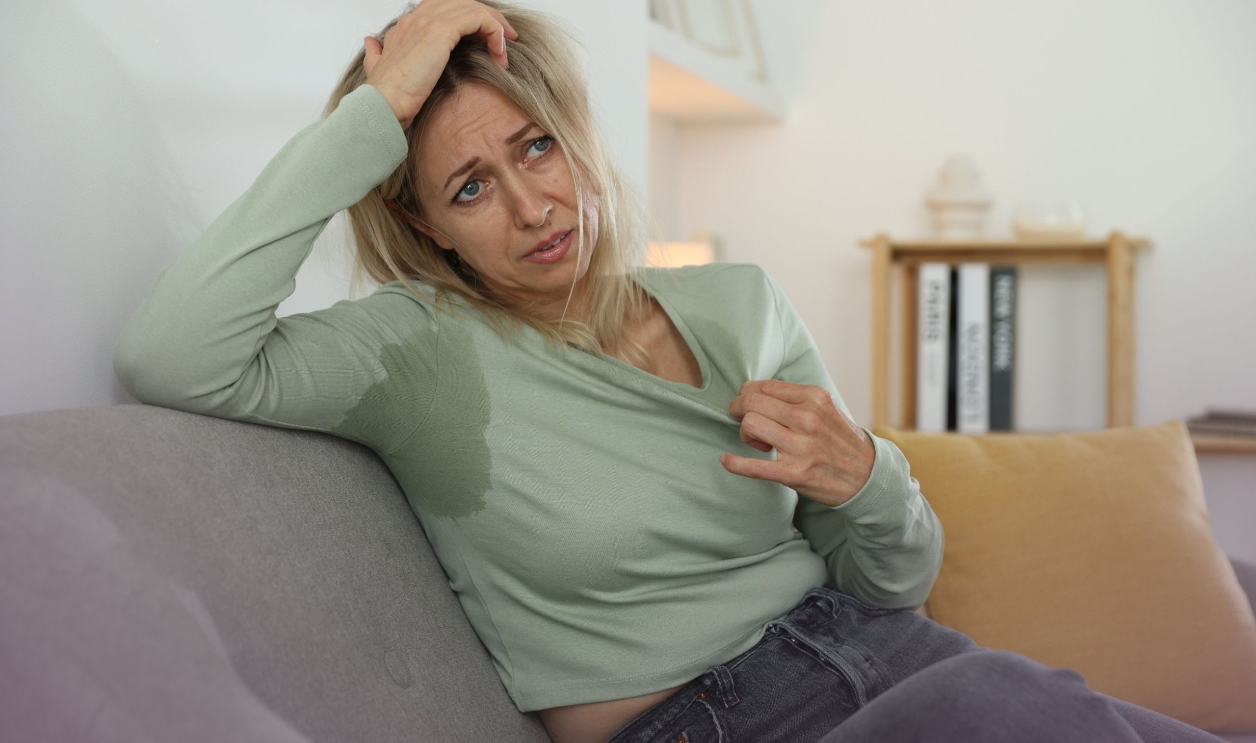 Frau mit Hitzewallungen sitzt erschöpft auf dem Sofa: Sie hat Schweißflecken unter den Achseln und zieht den Ausschnitt ihres Pullovers von sich, um mehr Luft an die schwitzende Haut zu lassen