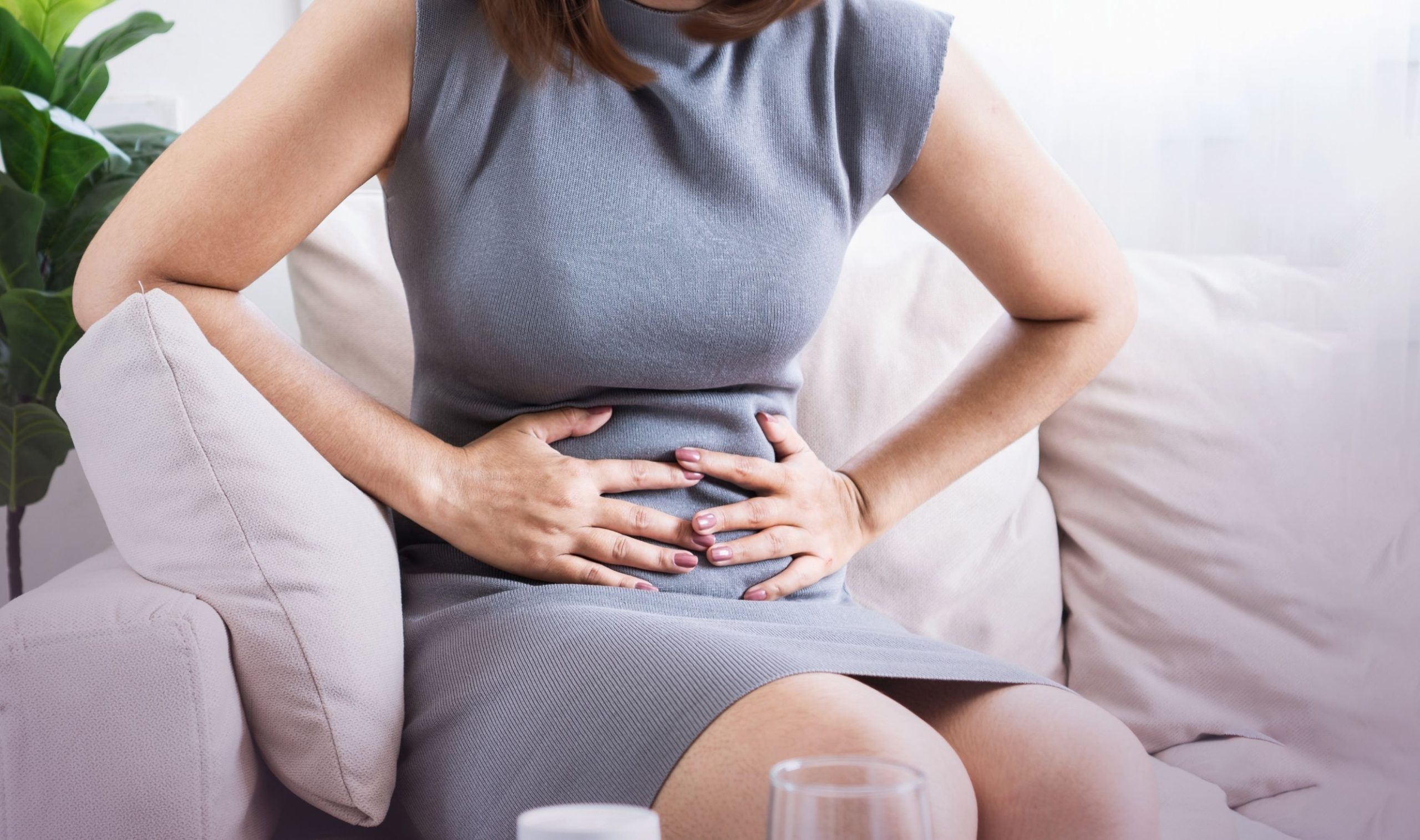 Frau mit Reizdarm Syndrom im Business-Kleid auf einem Sofa setzend, die Hände auf den schmerzenden Bauch gedrückt