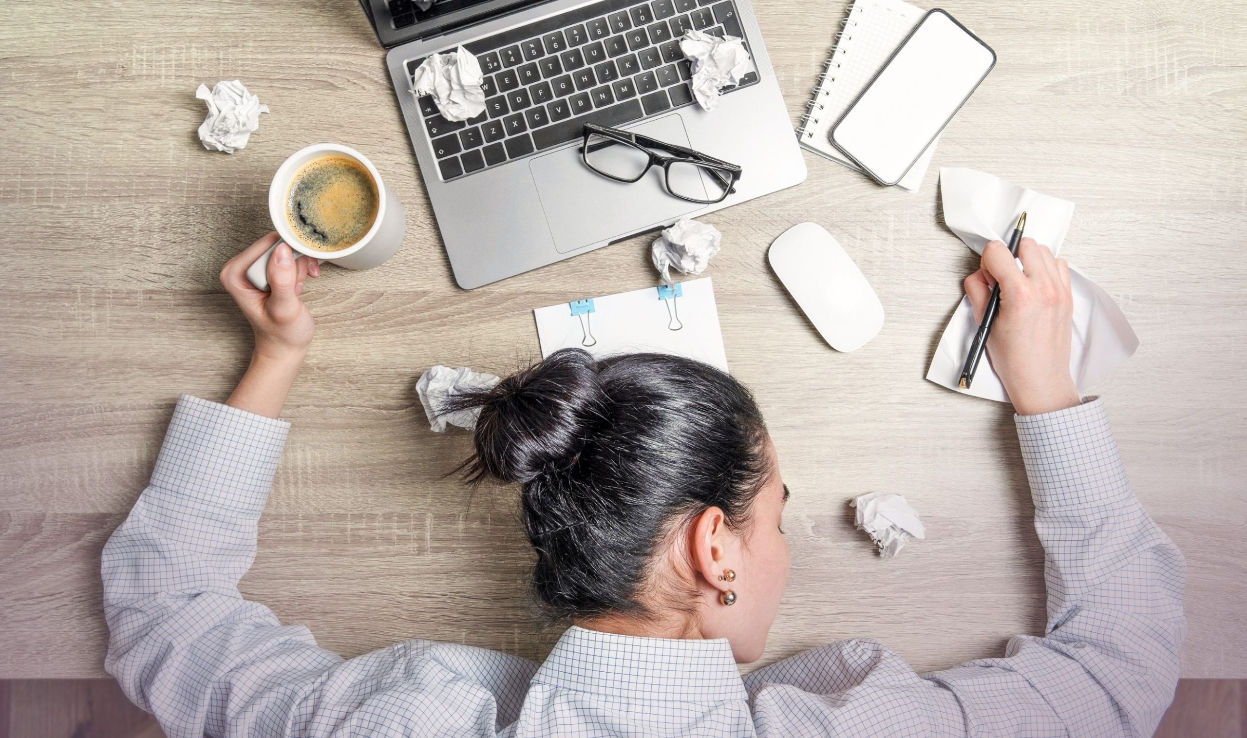 Frau, die unter Stress und Erschöpfung leidet, liegt mit dem Kopf auf dem überfüllten Schreibtisch mit Zetteln, Laptop und Kaffeetasse