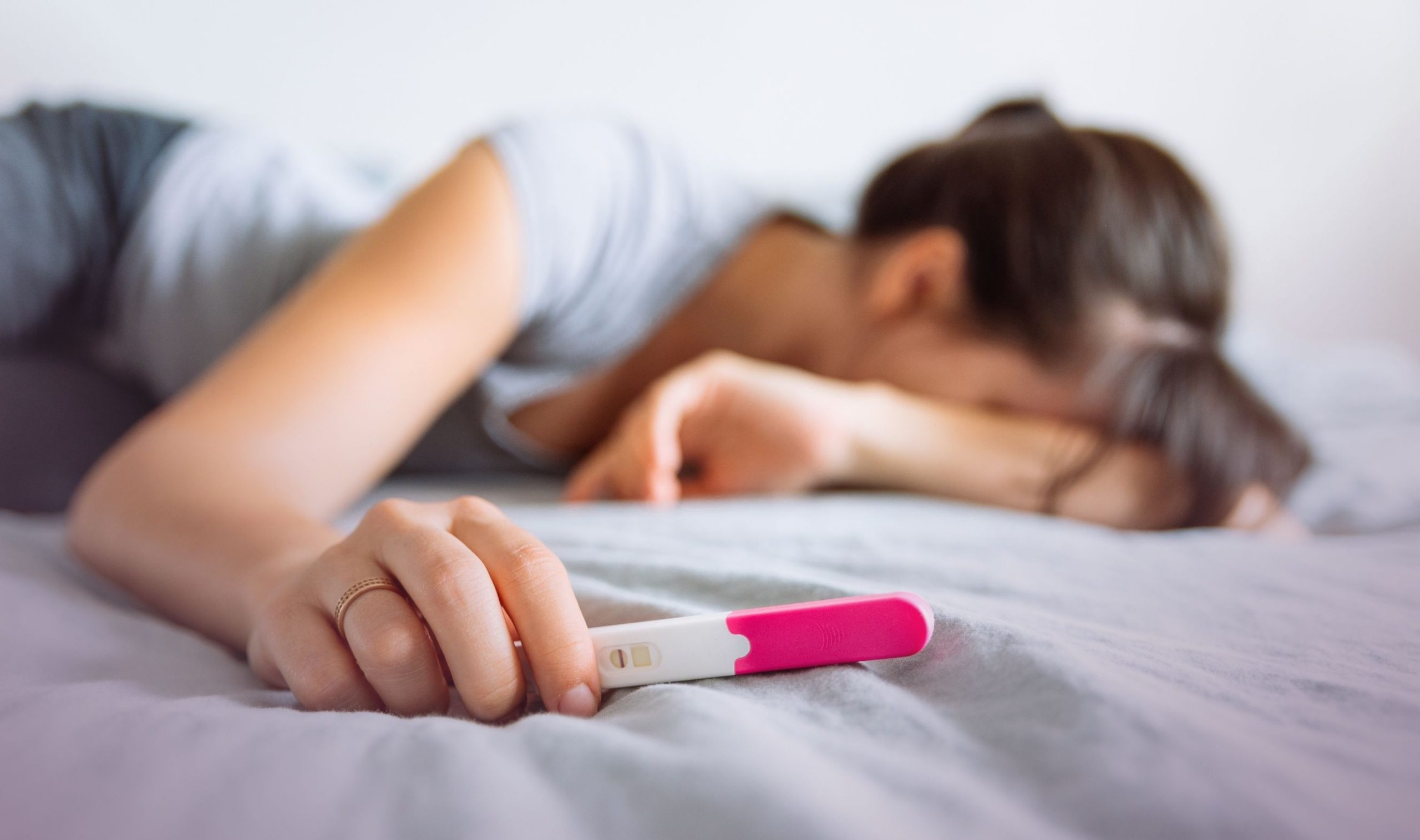Unerfüllter Kinderwunsch: Frau liegt auf dem Bauch auf einem Bett mit dem Kopf verzweifelt im Arm vergraben, in der rechten Hand einen negativen Schwangerschaftstest