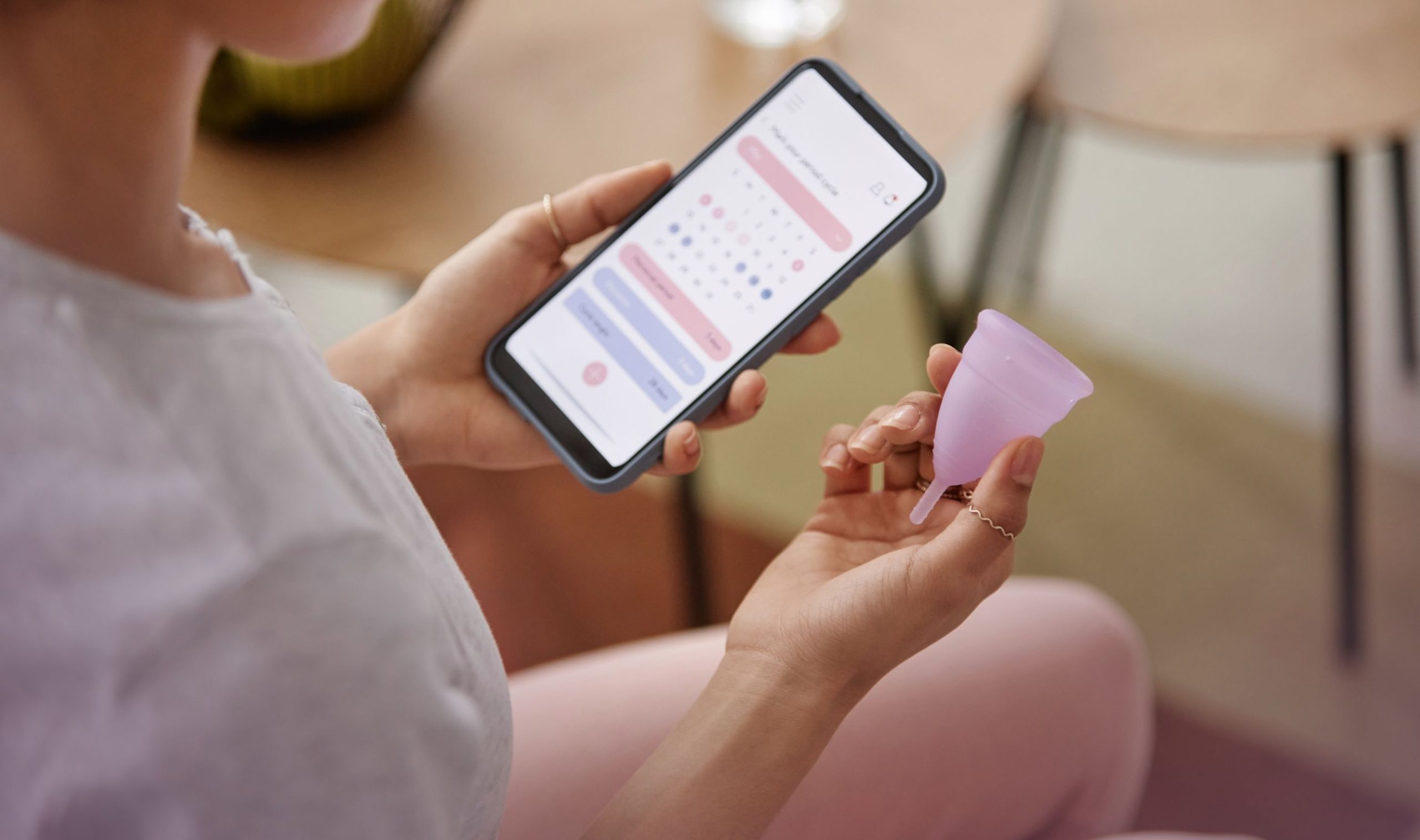 Frau mit Zyklusstörungen sitzt auf einem Stuhl, in einer Hand eine Menstruationstasse, in der anderen ihr Smartphone mit dem geöffneten Menstruationskalender